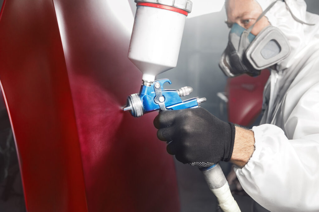 Professional spray painter in protective mask and clothes spraying car hood of vehicle part to red color using spray gun in paint chamber. Vehicle painting in a paint booth, restoration and repair.